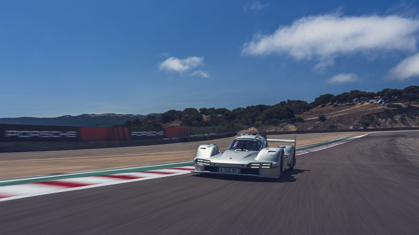 A white Porsche 963 RSP race car drives on a racetrack with hills and trees in the background under a blue sky, capturing the excitement of Car Week.