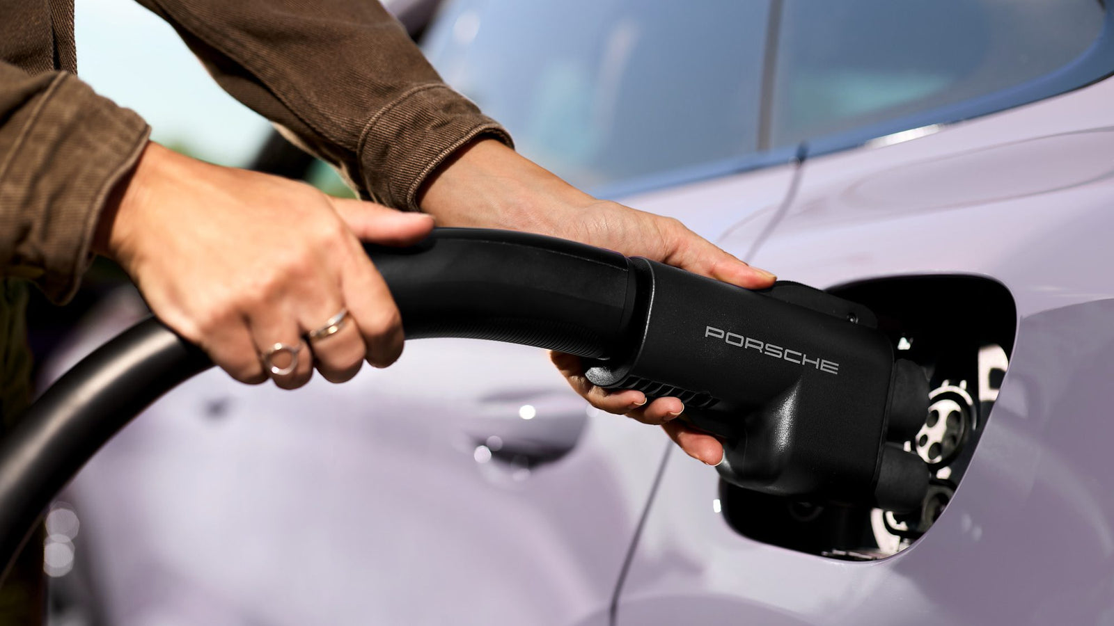 A person plugs a Porsche-branded electric vehicle charging cable into the charging port of a light-colored car, highlighting how Porsche EVs are now compatible with Tesla Superchargers across North America.