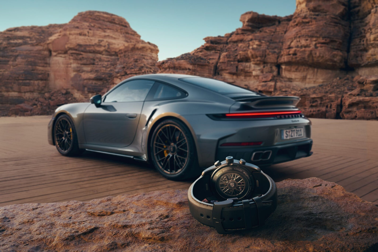 A luxury 911 Turbo S sports car is parked on a desert road with large rock formations in the background; a Porsche Design Custom-Built Chronograph wristwatch rests on a rock in the foreground.