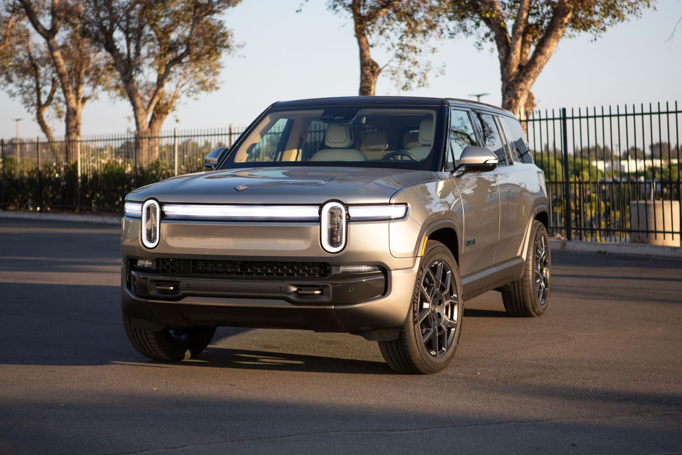 A silver Rivian R1S electric SUV is parked on a paved surface near a black metal fence and trees under daylight, ready to turn heads at Monterey Car Week.