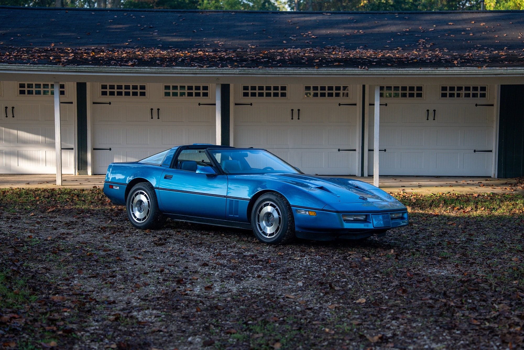 A blue Callaway Corvette, worthy of a Petrolicious Film Friday feature, is parked on a gravel surface in front of a row of white garage doors.
