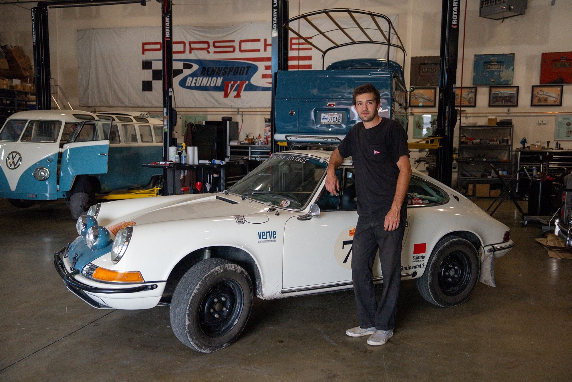 A man stands next to a vintage white Porsche sports car in a garage, with a blue Volkswagen van and various automotive tools in the background.