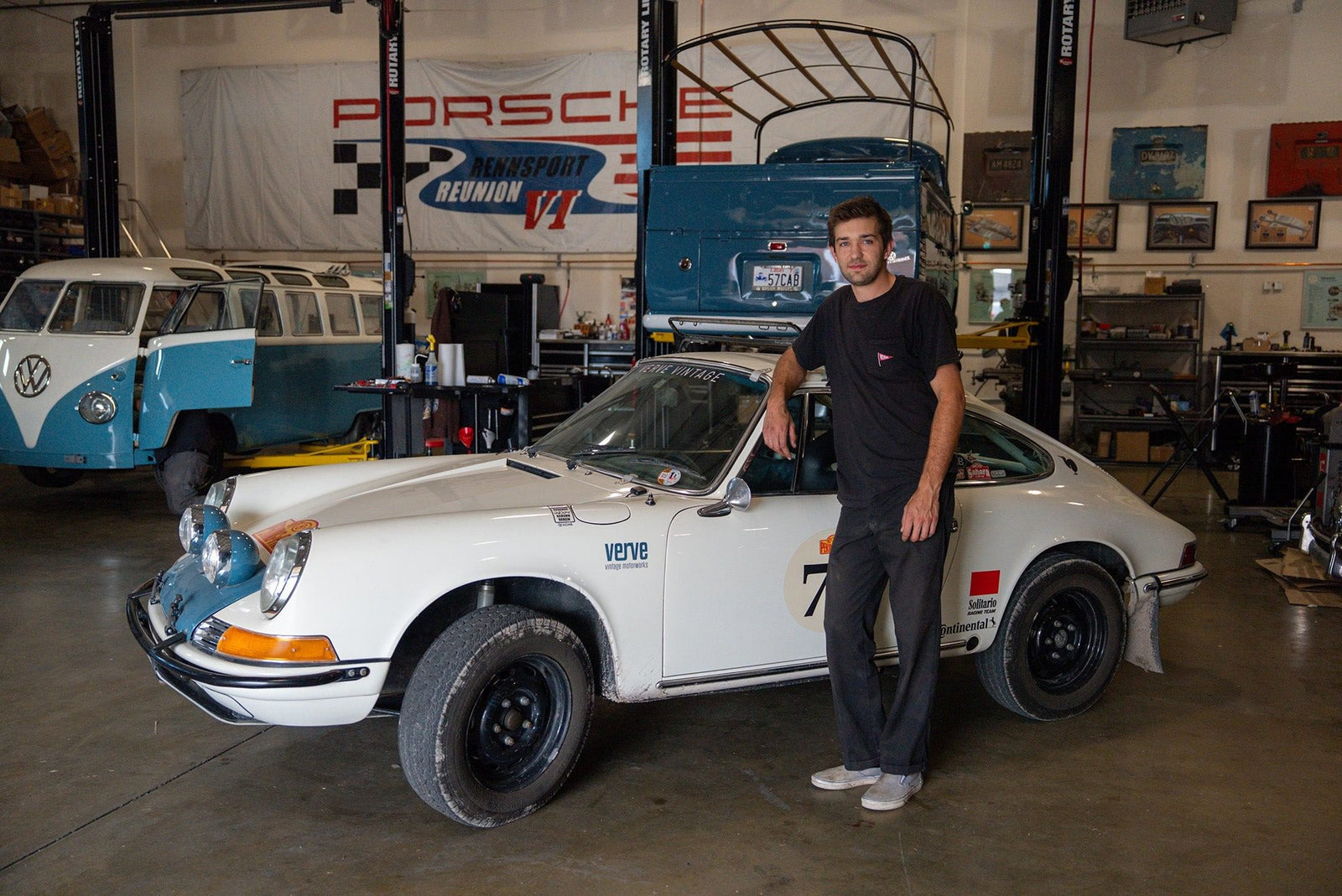 A man stands next to a vintage white Porsche sports car in a garage, with a blue Volkswagen van and various automotive tools in the background.