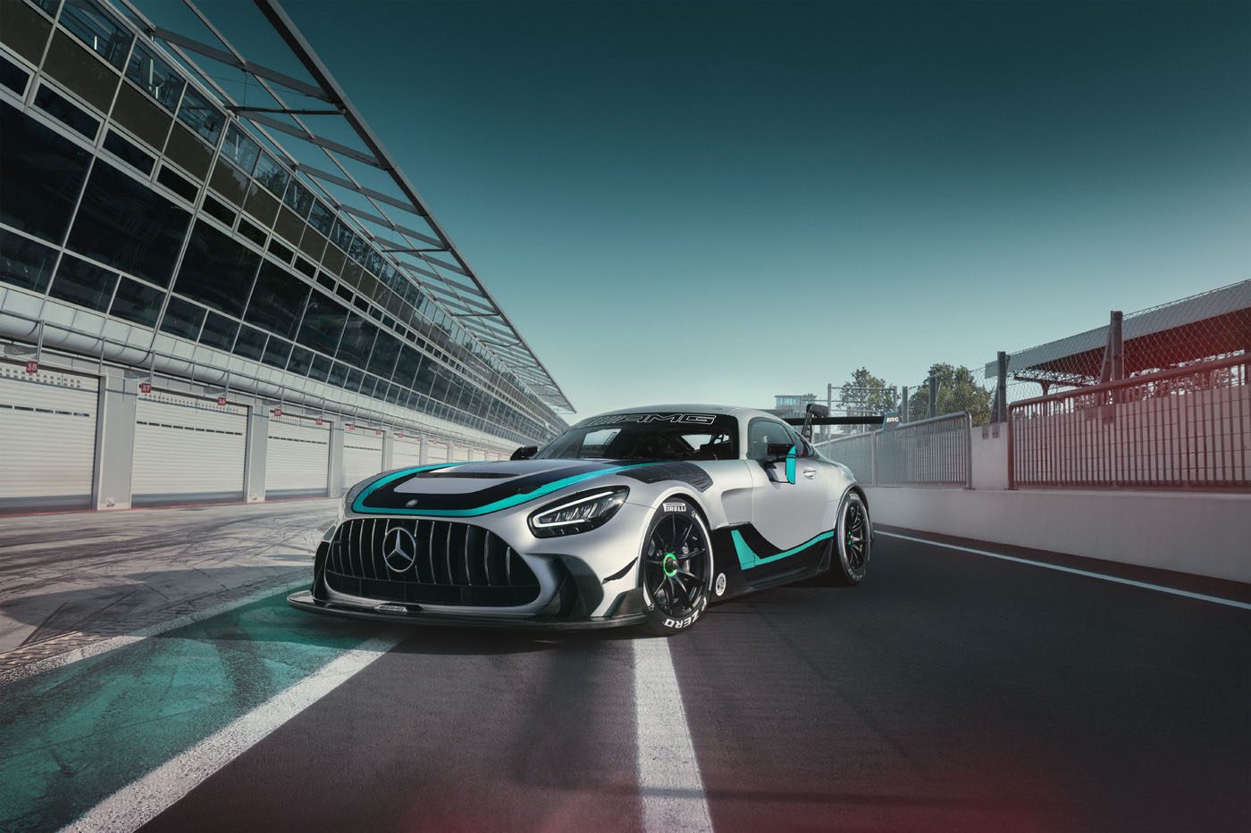 A silver Mercedes-AMG GT2 Edition sports car with turquoise accents is parked on a racetrack next to a pit lane under a clear sky.