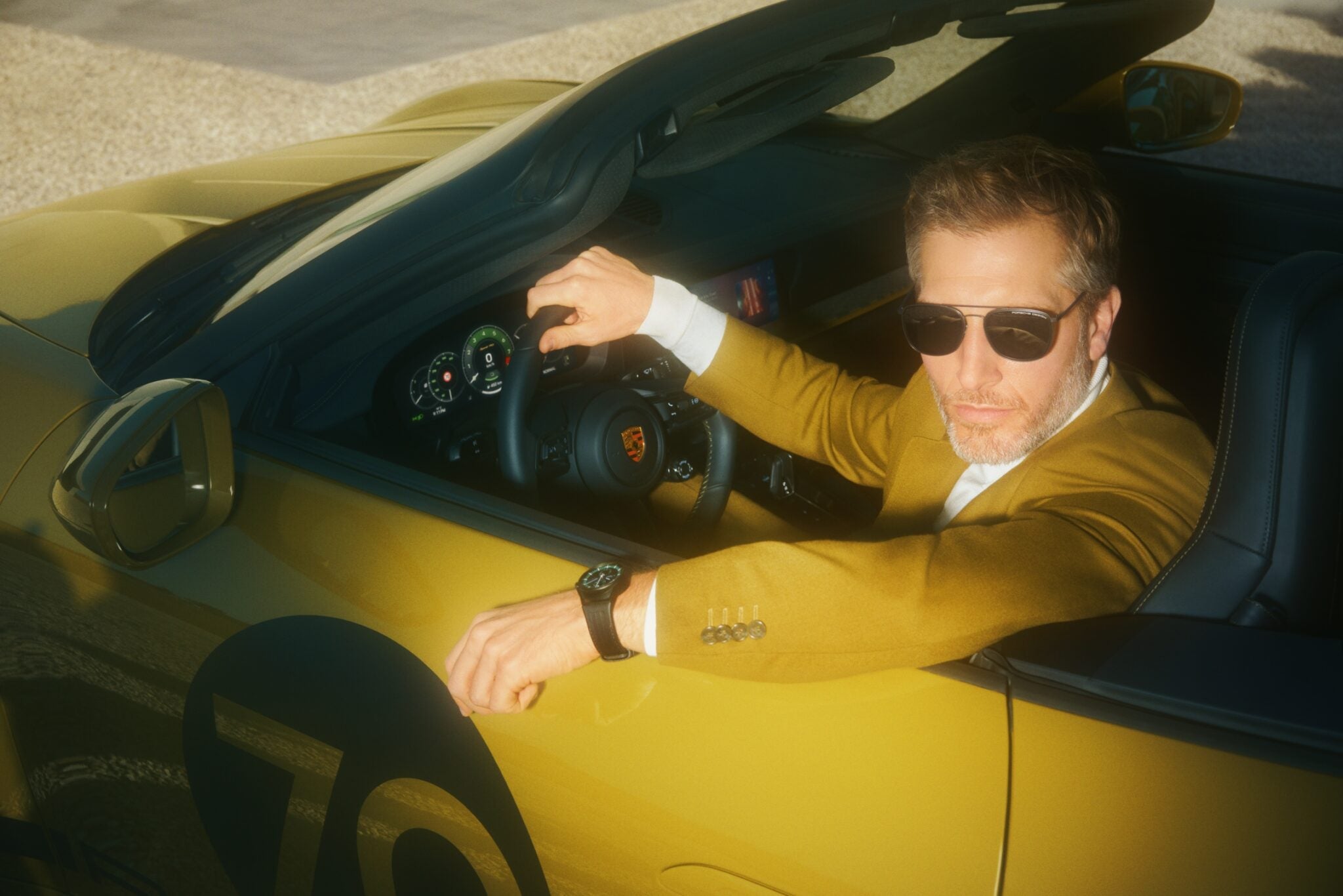 A man wearing sunglasses and a mustard-colored blazer sits in the driver’s seat of a gold convertible sports car, showcasing his Owner-Exclusive style and glancing toward the camera—his Porsche Design Chronograph 911 visible on his wrist.