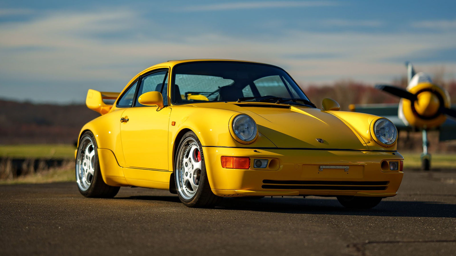 A yellow Porsche 911 Turbo gleams on the tarmac, poised beside a sleek airplane under the expansive blue sky—a scene that echoes the elegance often celebrated at Broad Arrow Auctions.