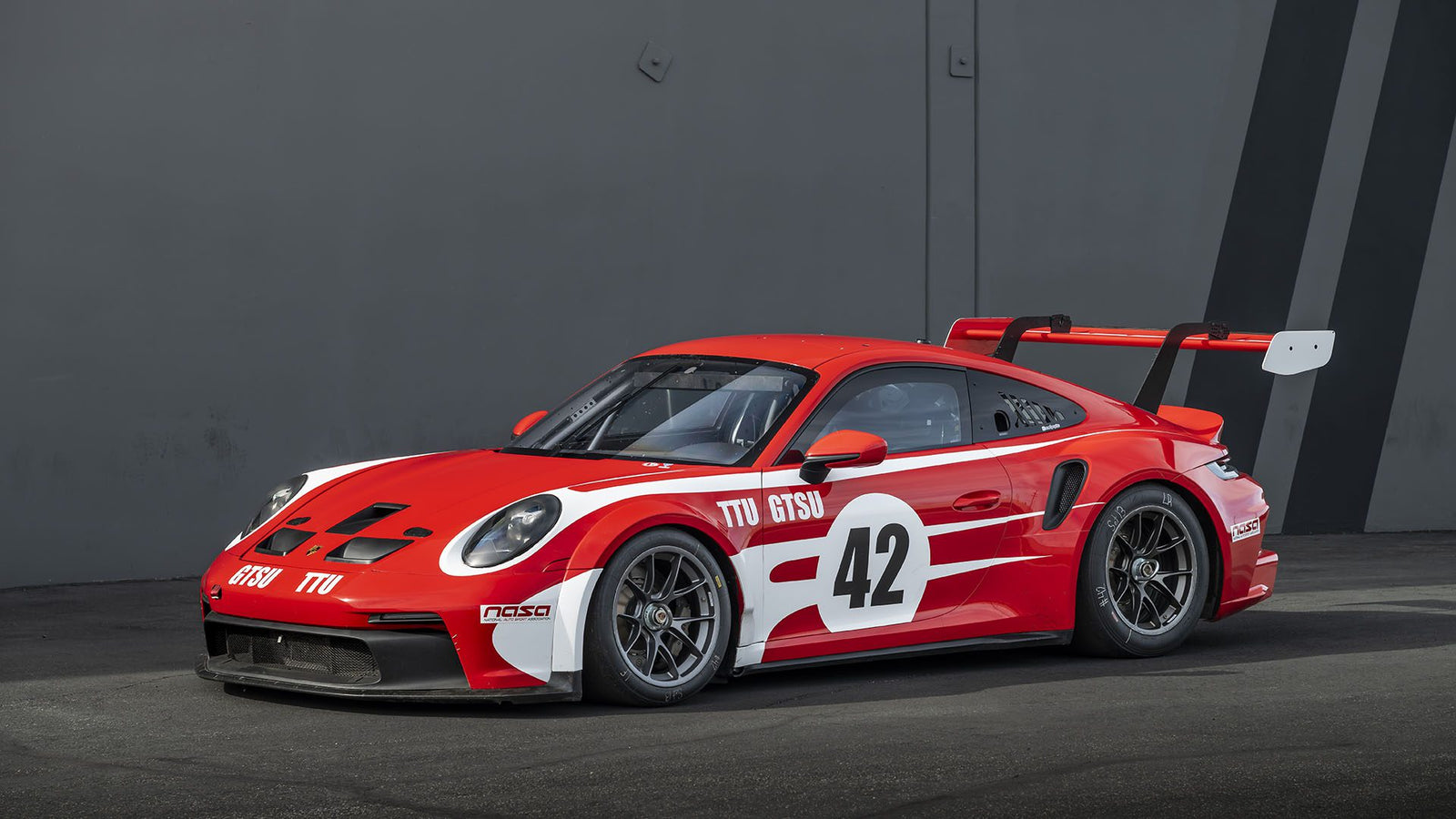 A red and white Porsche 911 GT3 with the number 42 on the side, featuring a large rear spoiler, is parked gracefully on the gray pavement. Don’t miss its showcase at Broad Arrow Auctions.