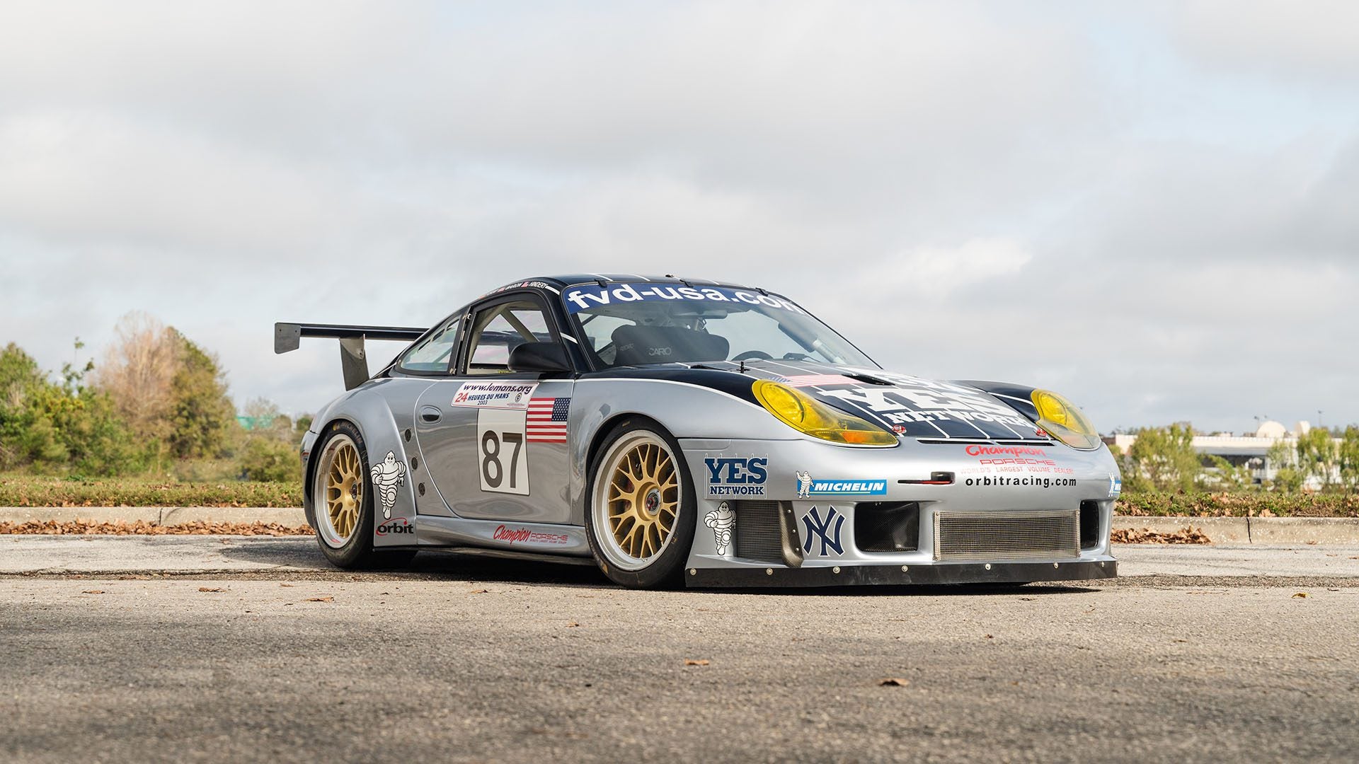 A sleek silver Porsche 911 GT3 RS, boasting the number 87 and adorned with Orbit Racing sponsor logos, is parked on a street lined with trees and buildings.