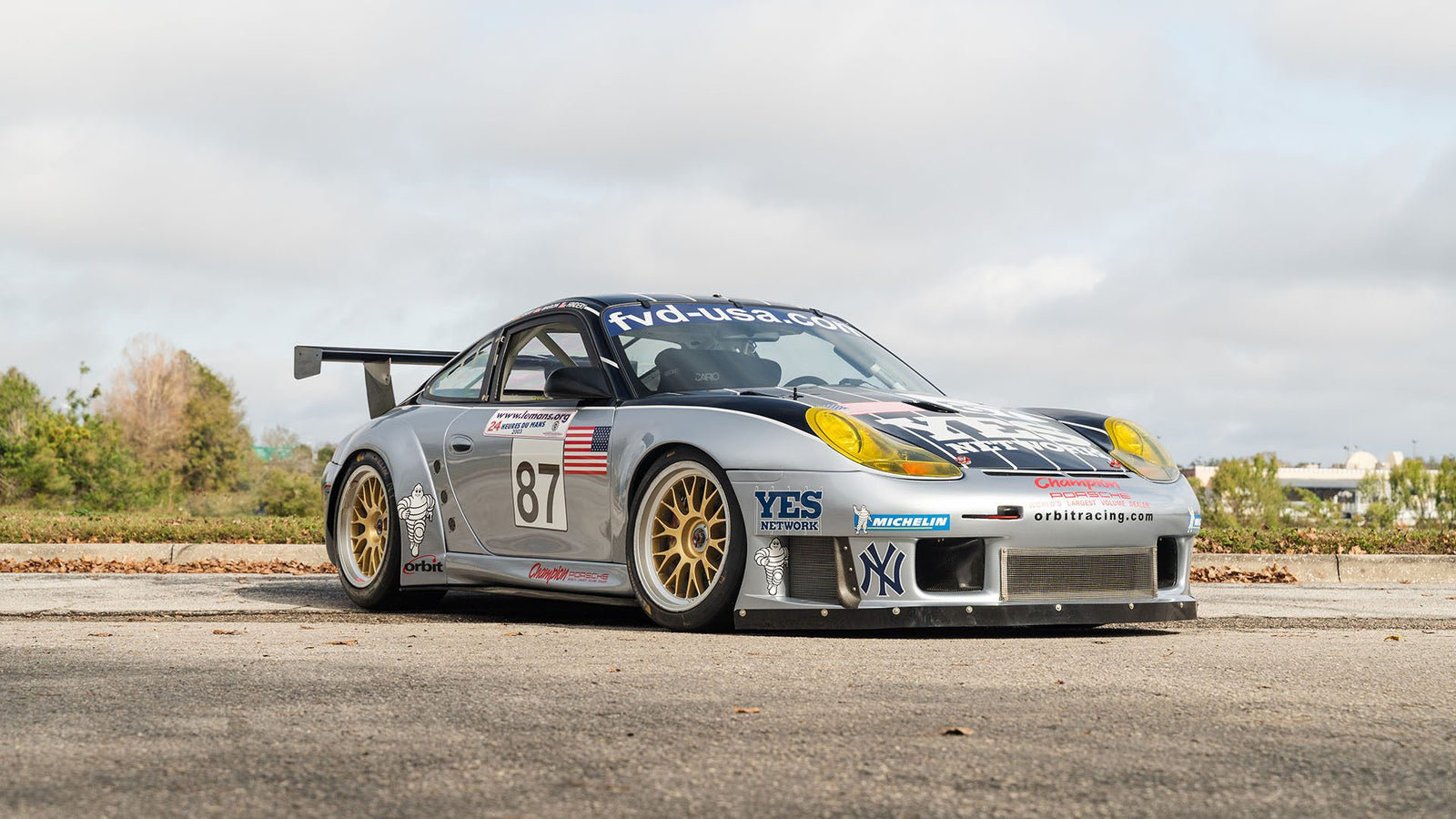 A sleek silver Porsche 911 GT3 RS, boasting the number 87 and adorned with Orbit Racing sponsor logos, is parked on a street lined with trees and buildings.