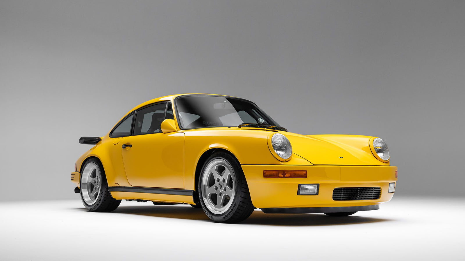 A 1989 RUF CTR Yellowbird, with its iconic yellow finish, two doors, circular headlights, and wide tires gleams under the bright studio lights. It's a striking piece of automotive history that would be a highlight at Broad Arrow Auctions.