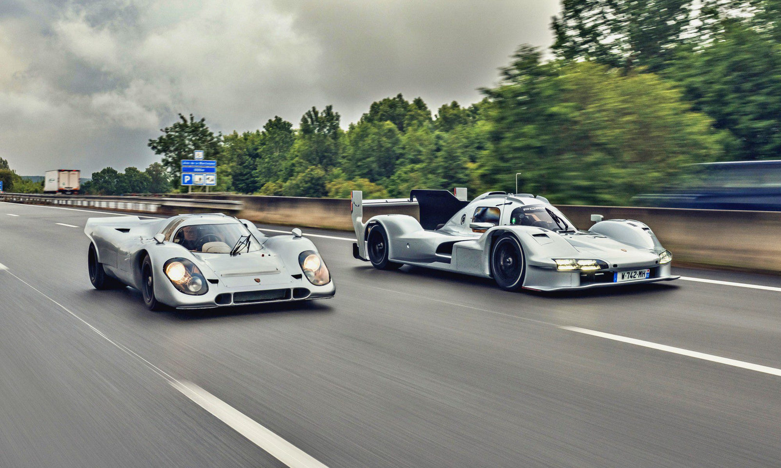Two white Porsche 963 race cars, part of a Penske Tribute, drive side by side on a highway with trees and road signs in the background.
