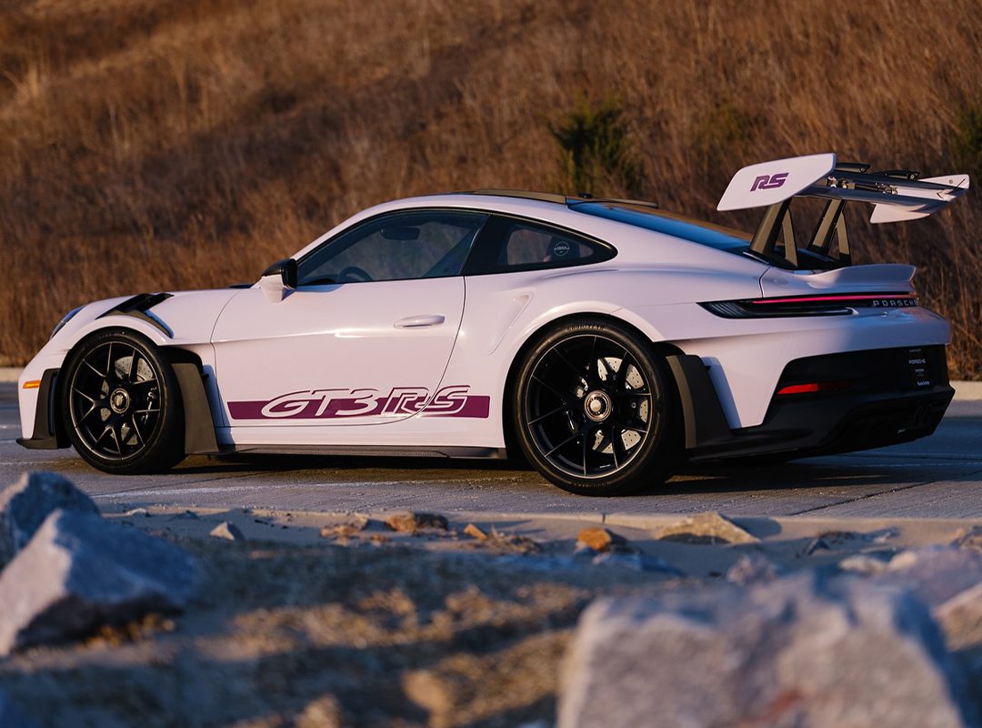 A sleek white 911 GT3 RS with a PTS Moongem finish flaunts its "GT3 RS" decal and large rear wing, resting on black wheels. It's parked stylishly on a dirt road, framed by dry grass, as if fresh from the Porsche Exchange showroom.