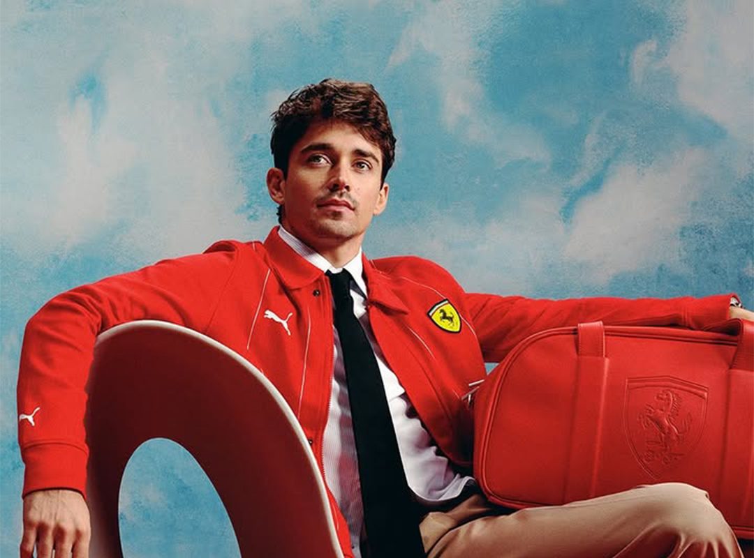 A person in a red jacket with a Ferrari logo sits on a modern chair, holding a matching red bag from the latest PUMA Motorsports collection. The background is a blue sky with clouds.