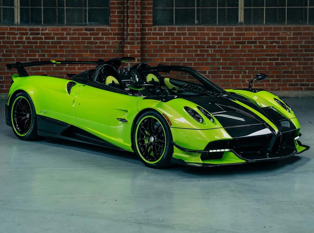 A bright green and black Pagani hypercar is parked indoors in front of a brick wall and large windows.