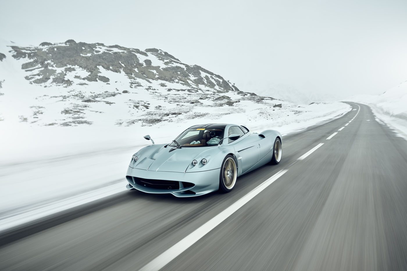 A silver Pagani hypercar drives on an empty, snow-lined mountain road under an overcast sky.
