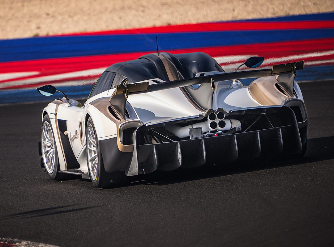 A silver Pagani Huayra R Evo with a large rear wing and distinctive exhaust pipes drives on a racetrack with blue, red, and white curbs, capturing the spirit of the Milano Monza Motor Show.