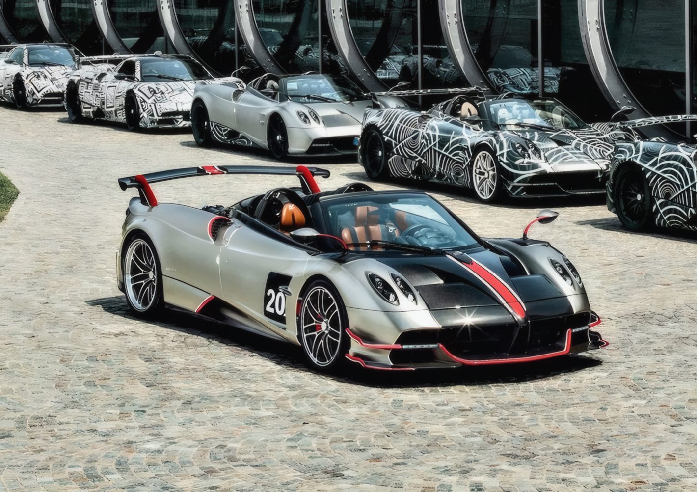 A silver and black sports car with red accents is parked on a cobblestone driveway, surrounded by similar cars—possibly stars like those Pagani will showcase at the upcoming 9th Historic Minardi Day in Imola.