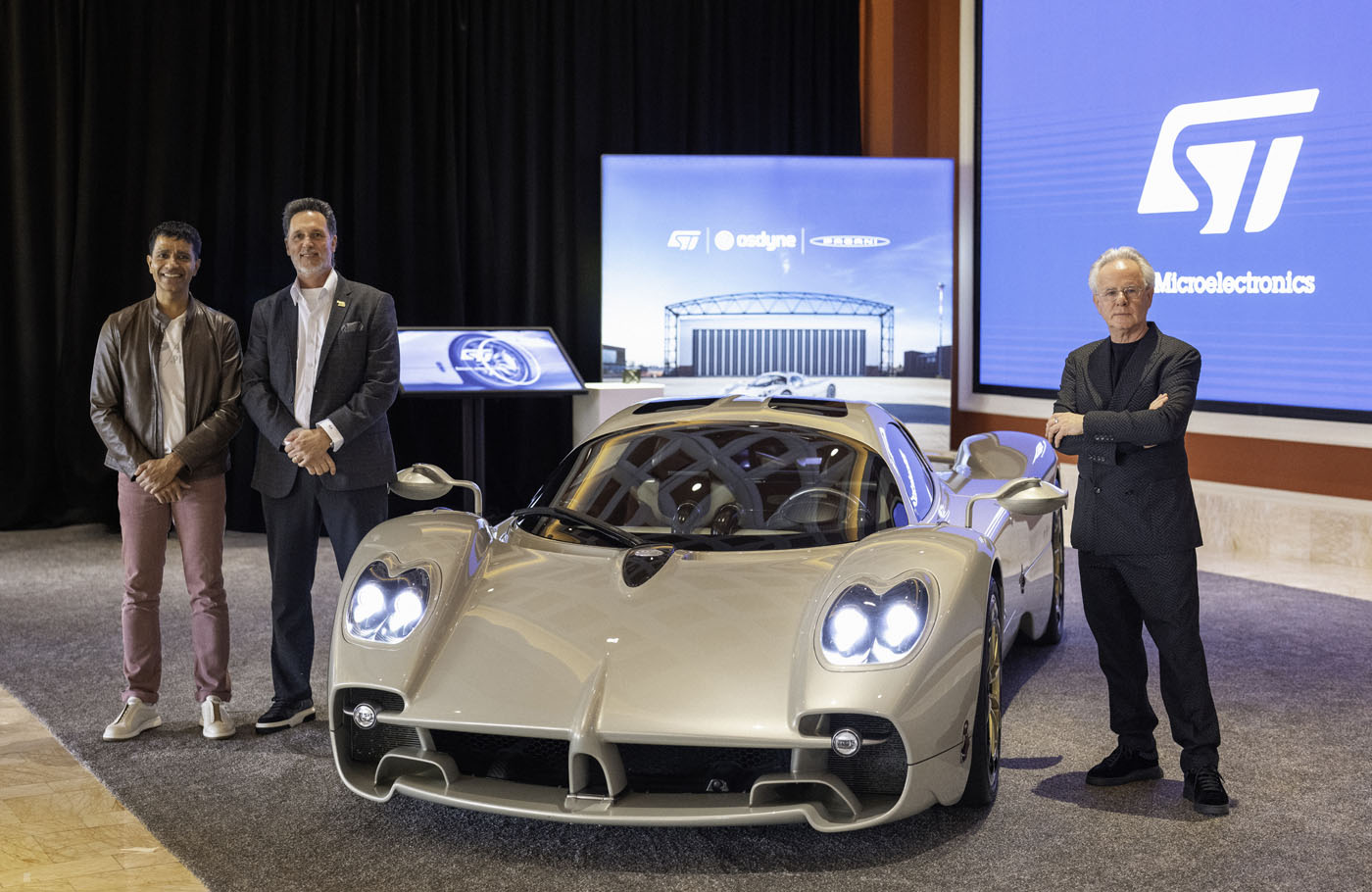 Three men stand beside a white Pagani hypercar at an indoor event, with display screens in the background showcasing STMicroelectronics technology branding.