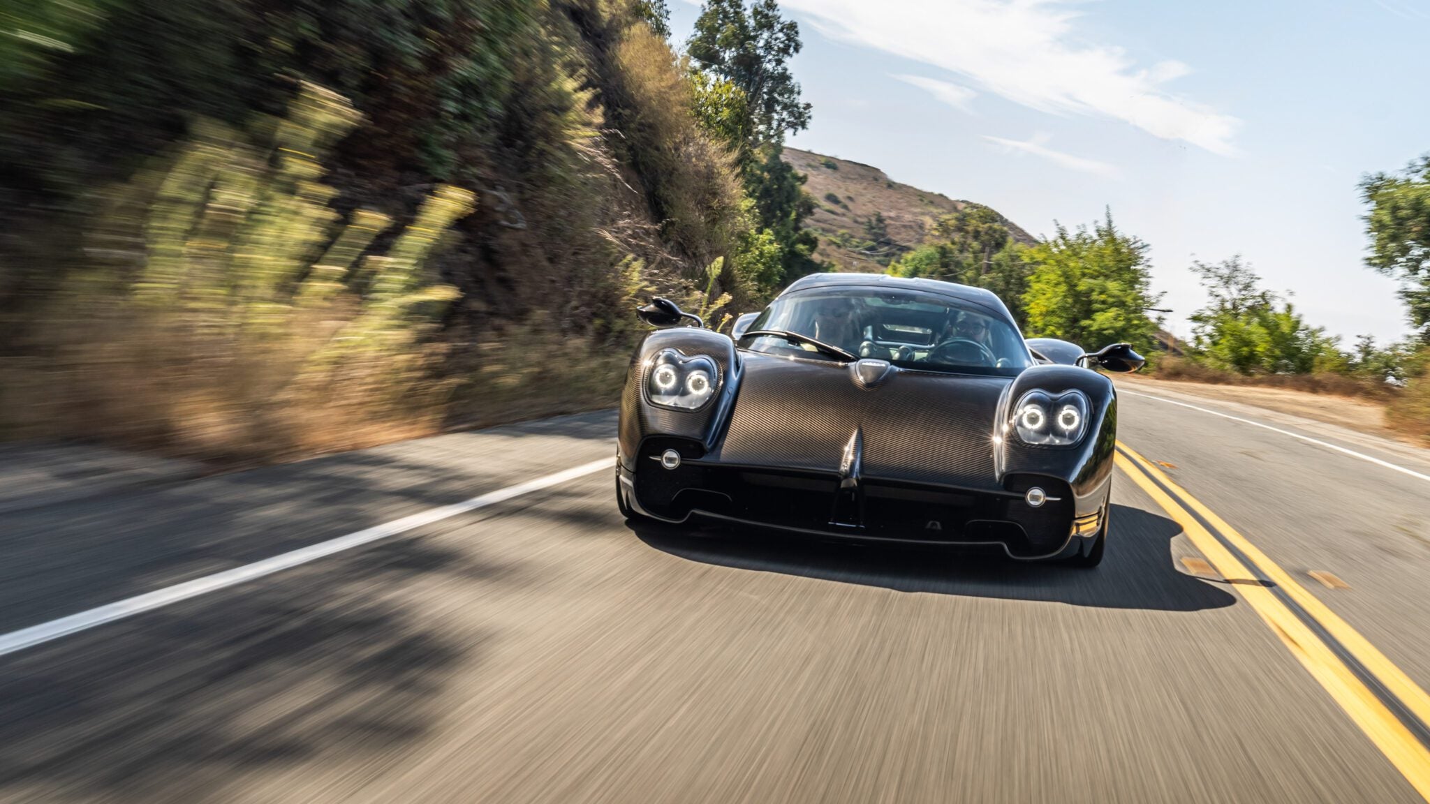 A sleek Pagani Utopia sports car glides along a scenic, winding road, embraced by lush greenery and rolling hills beneath a clear sky.