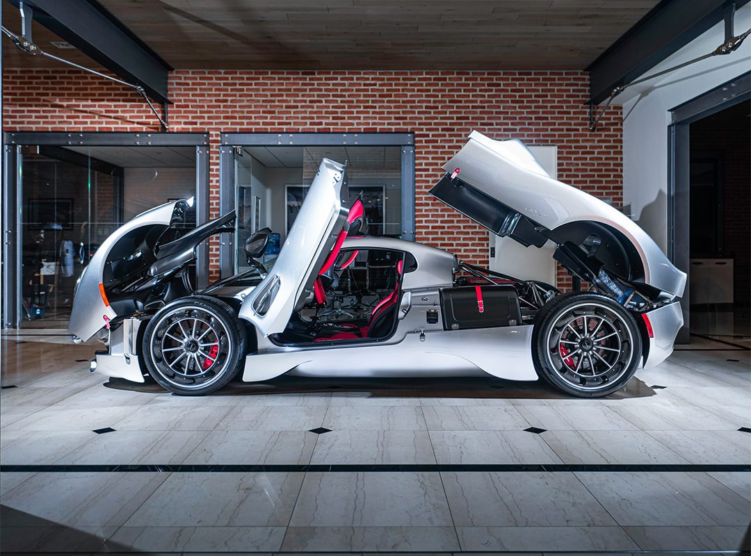 A sleek Pagani Utopia with its doors and panels open stands proudly, awaiting its new owner. Nestled indoors on a tiled floor against a brick wall backdrop, this masterpiece is every car enthusiast’s dream, epitomizing the excellence of Miller Motorcars.