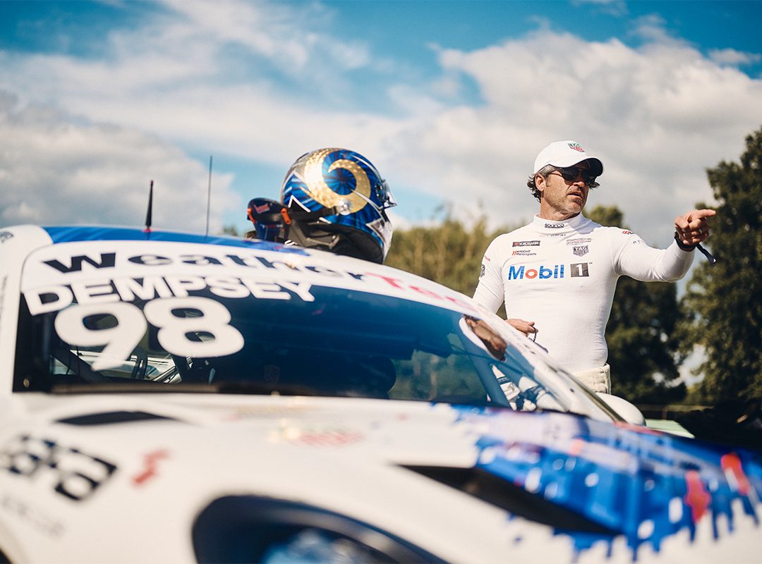 Race car driver in a white suit stands beside car number 98 with "Dempsey" on the windshield, marking a star turn in his racing career. A helmet rests on the car. The driver points, wearing sunglasses and a cap.