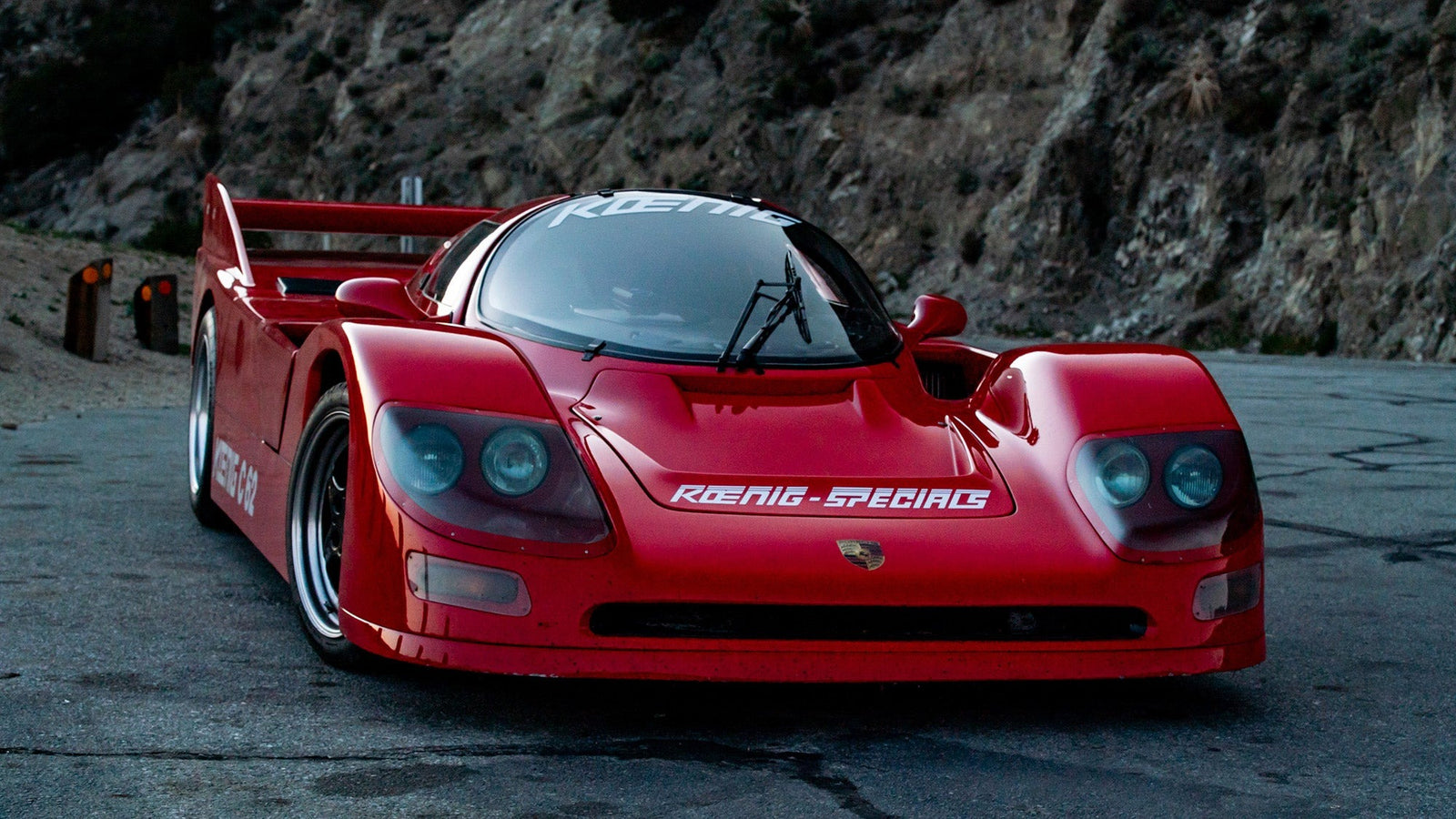 A red sports car with the "Koenig-Special" label proudly displayed on its hood, the C-62 Special is parked on a winding mountain road. Rugged rocky cliffs stand tall in the background, creating a scene fit for a Petrolicious feature.
