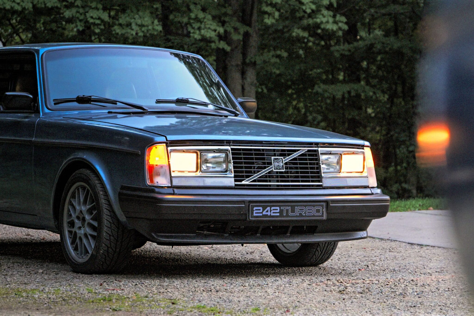 A classic Volvo 242 Turbo, reminiscent of its Group A Homologation days, with headlights on, parked on a gravel path against a backdrop of trees—a scene straight out of a Petrolicious story.