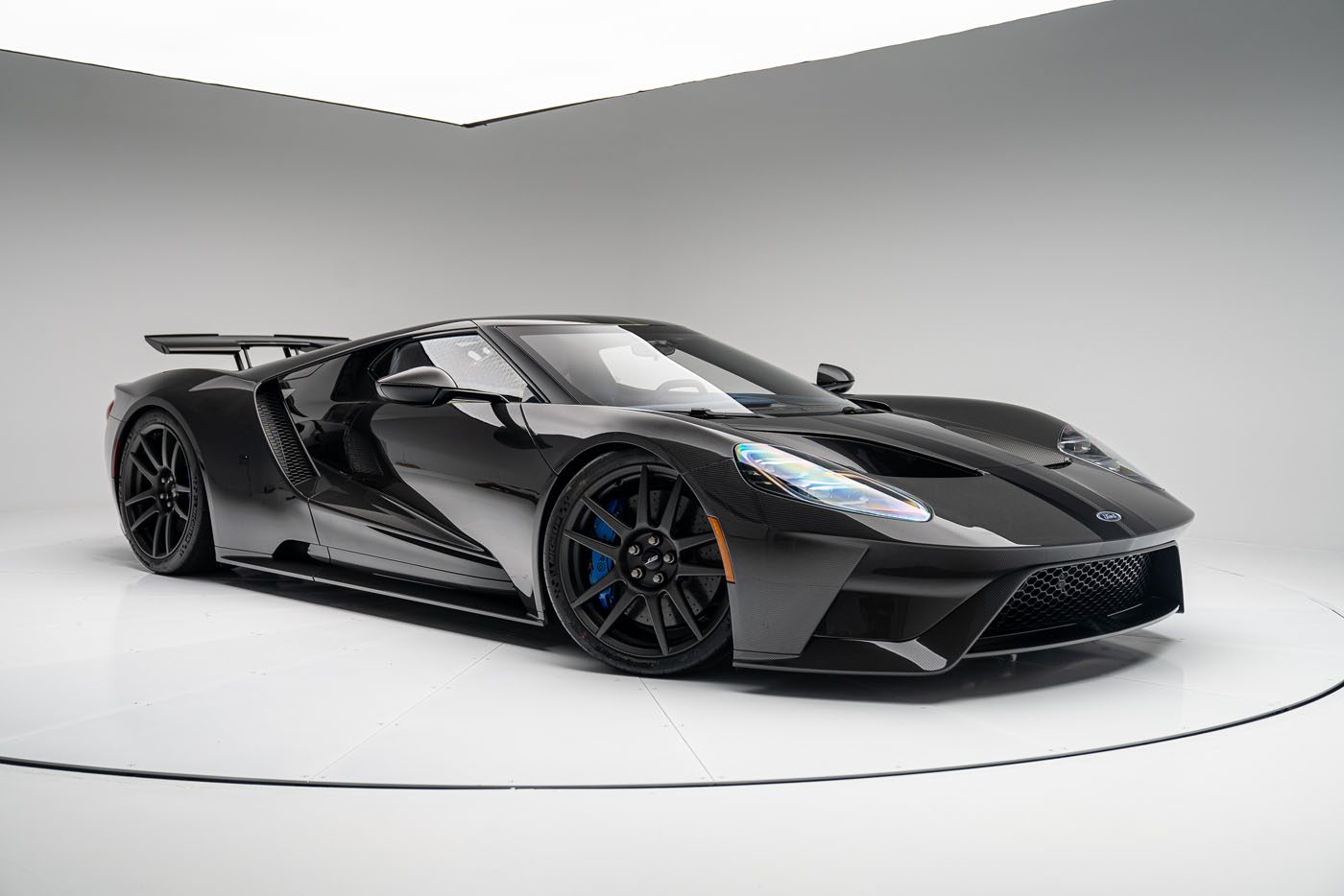 A sleek black Ford GT sports car with a low, aerodynamic design, large wheels, and a rear spoiler is parked in a white studio setting. The Liquid Carbon Edition subtly flaunts ghosted stripes that add an extra layer of intrigue to its flawless finish.
