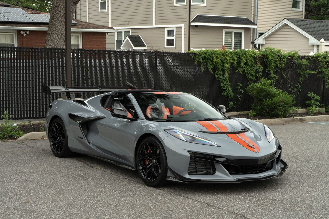 A gray 2025 Chevrolet Corvette ZR1 with black wheels and orange racing stripes is parked on a street in front of a black fence and houses, looking like it’s ready for sale to an eager new owner.