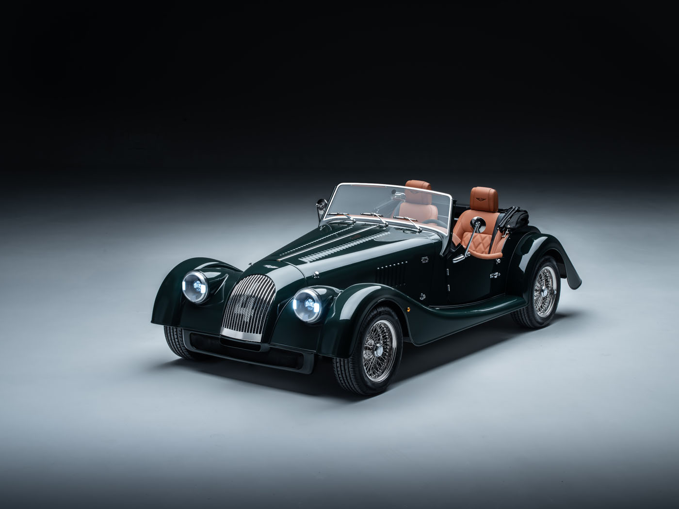A classic green Morgan convertible car with brown leather seats, wire wheels, and a vintage design, the Plus Four is elegantly displayed in a studio setting.