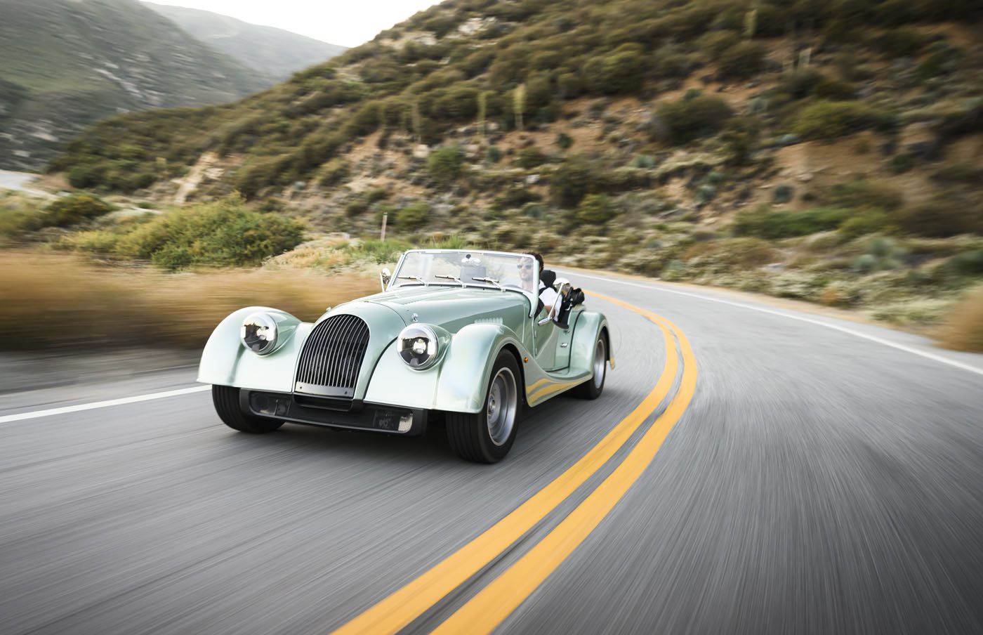 A classic mint green Morgan Plus Four convertible car drives along a winding mountain road lined with vegetation, celebrating the 75th Anniversary of this iconic model in the US.