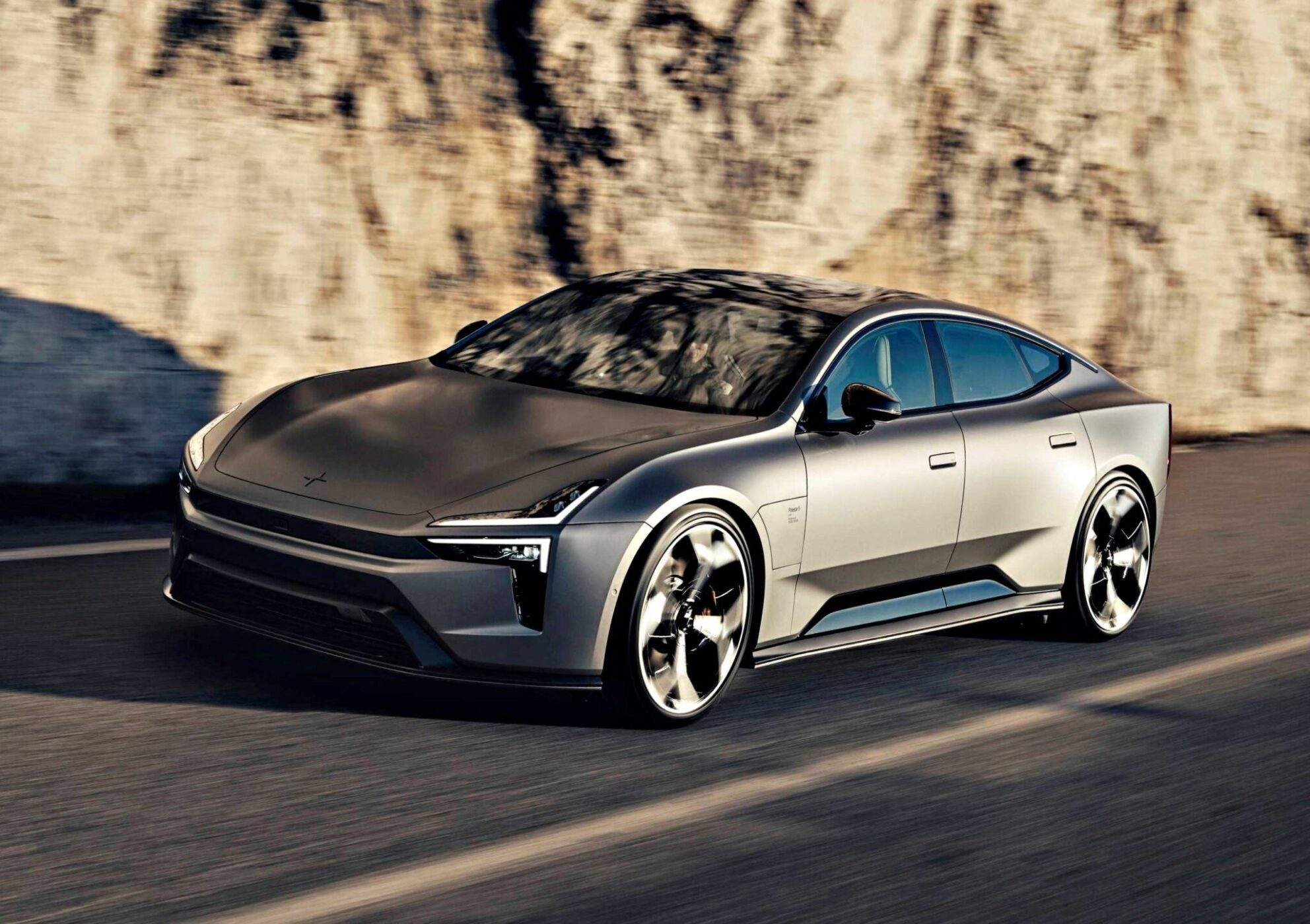 A sleek, silver electric sedan with aerodynamic lines—possibly the Polestar 5, which debuts at IAA 2025 as the brand’s first true flagship EV GT—drives along a road beside a rocky wall.