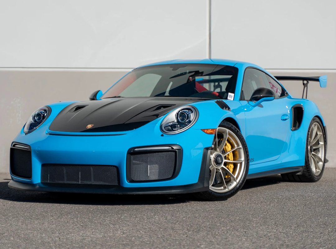 A blue 991 Porsche 911 GT2 RS with a black hood and large rear wing, known for its skyrocketing value, is parked on a paved surface in front of a plain, light-colored wall.