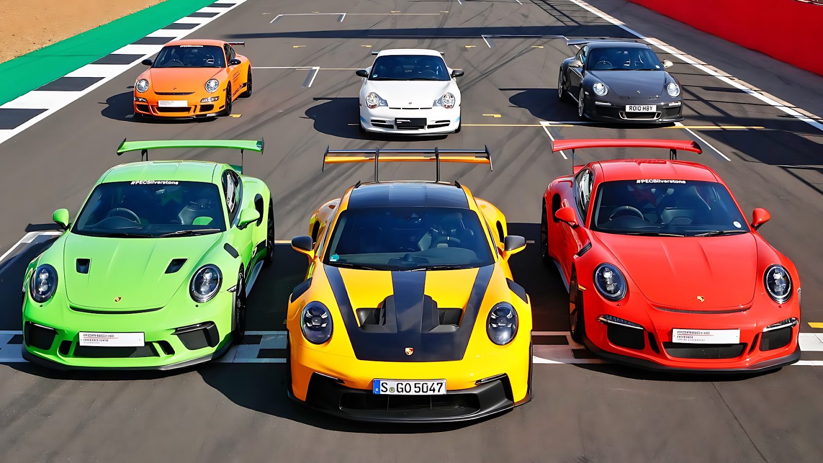 Six Porsche sports cars, including the iconic Porsche 911 GT3 RS in vibrant colors, are parked on a racetrack in two neat rows, all facing forward.