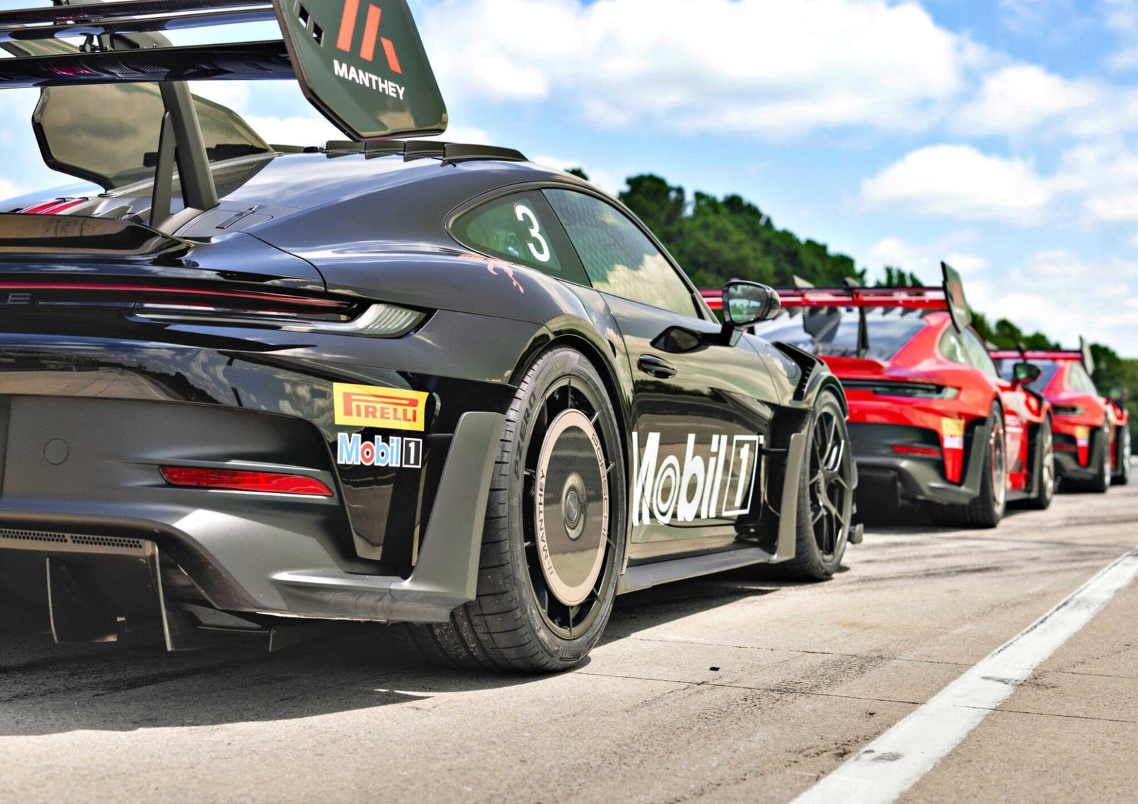 Three race cars with large rear wings, including a Porsche 911 GT3 RS Manthey Package now offered at Porsche Track Experience in Alabama, are lined up on a track, viewed from the rear, featuring Mobil 1 and Pirelli branding.