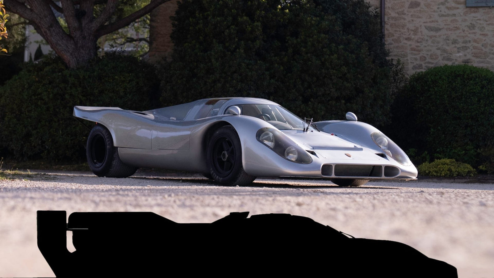 A vintage silver race car, reminiscent of a street-legal 917, is parked on a gravel driveway, with trees and a stone building in the background. The silhouette of a sports car celebrates Porsche 50 years at the bottom of the image.