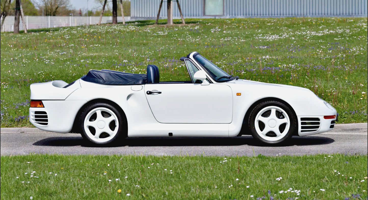 A white Porsche 959 Speedster with the top down is parked on a paved road next to a grassy field with scattered flowers, ready to turn heads at the RM Sotheby's Milan Auction.