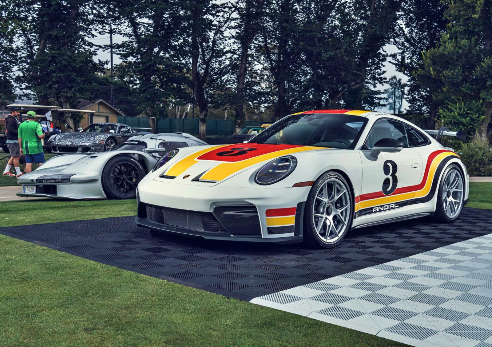 A white Porsche 911 GT3 ANDIAL Wrap with red and yellow racing stripes and the number 3 on the hood is displayed outdoors beside another silver sports car at a show where Penske’s 963 RSP headlines Monterey.