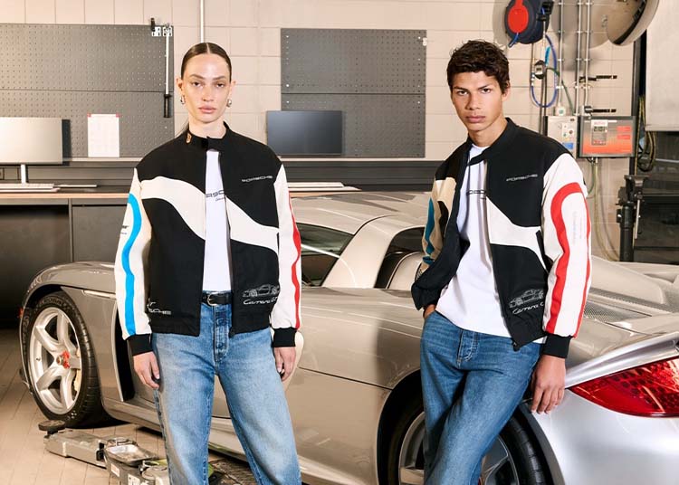 Two people in matching black, white, and colored racing jackets and blue jeans stand in front of a silver Porsche Carrera GT, part of an Exclusive Collection, in a garage setting.