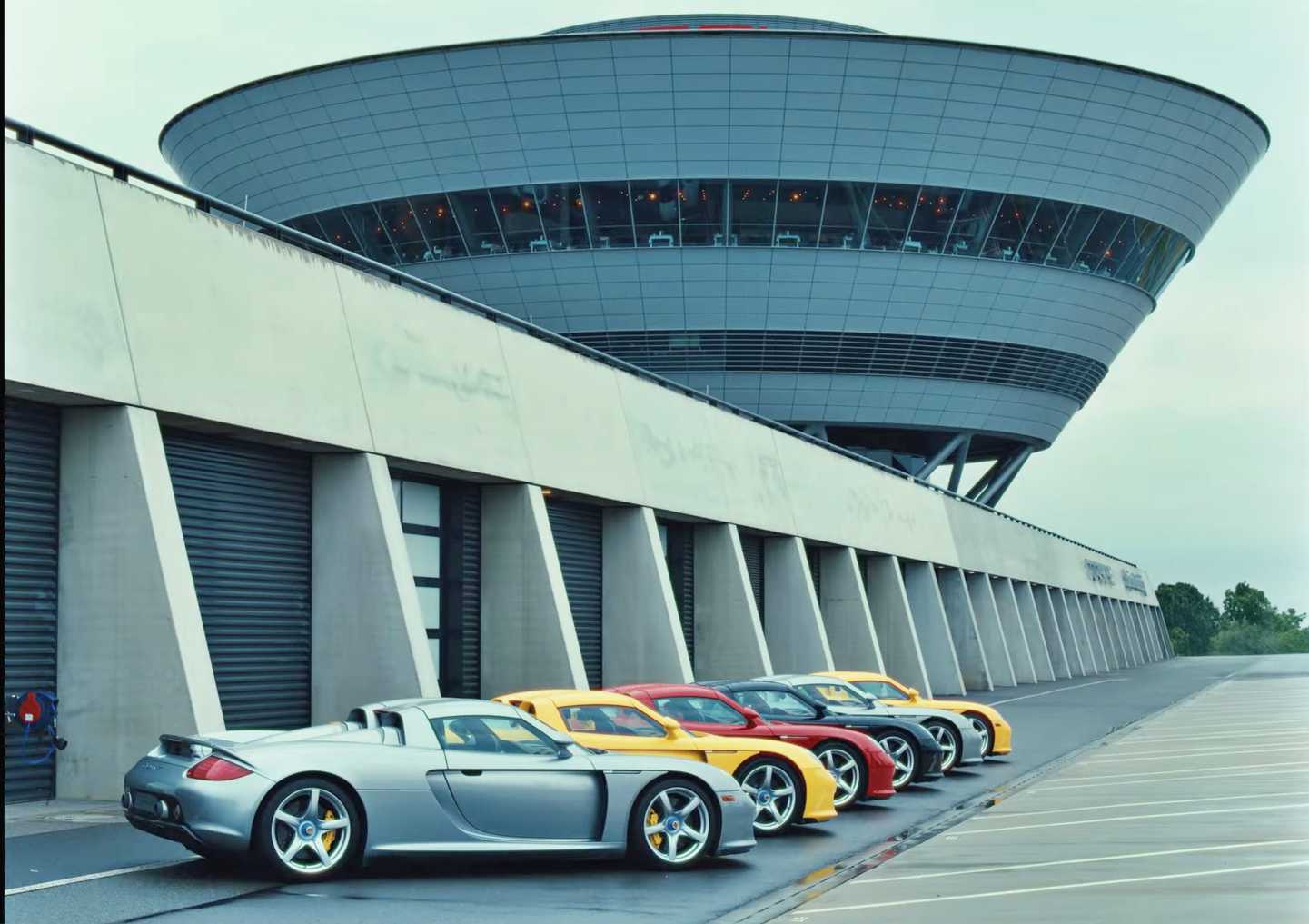 Five Porsche sports cars, including a striking Porsche Carrera GT, are parked in a row outside a modern, circular glass and steel building on an overcast day, capturing the spirit of a Carrera GT anniversary.