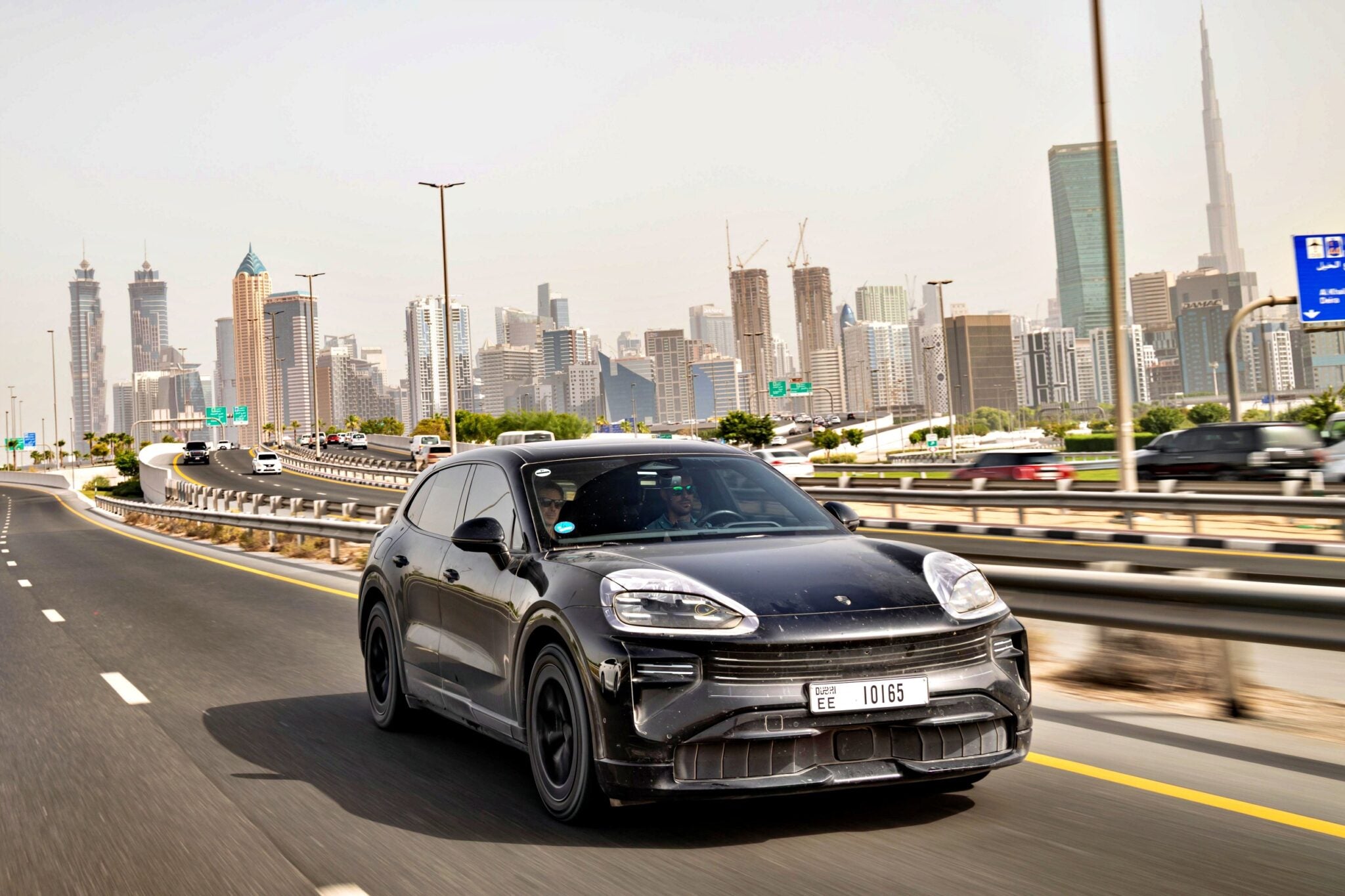 A black Porsche All-Electric Cayenne drives on a multi-lane highway in an urban area, surrounded by modern skyscrapers and construction cranes in the background.
