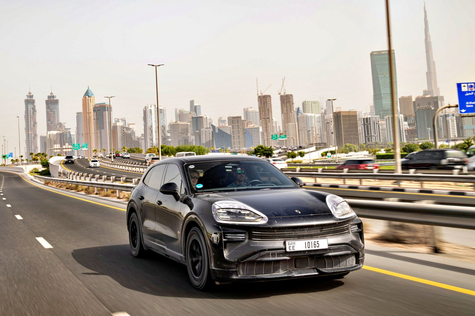A black Porsche All-Electric Cayenne drives on a multi-lane highway in an urban area, surrounded by modern skyscrapers and construction cranes in the background.