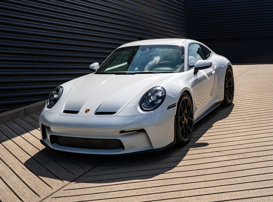 A white Porsche sports car from Porsche Centre Langley is parked on a paved, striped surface next to a dark metal wall under daylight—an eye-catching dealer spotlight for exotics for sale.