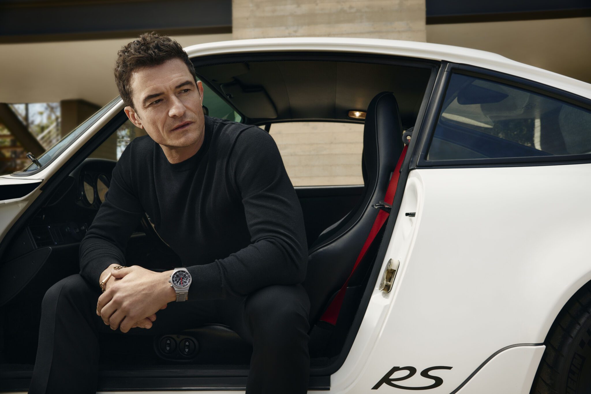 A man in a black outfit, reminiscent of Orlando Bloom’s sleek style, sits in the open doorway of a white car with "RS" lettering, looking to the side with a neutral expression.