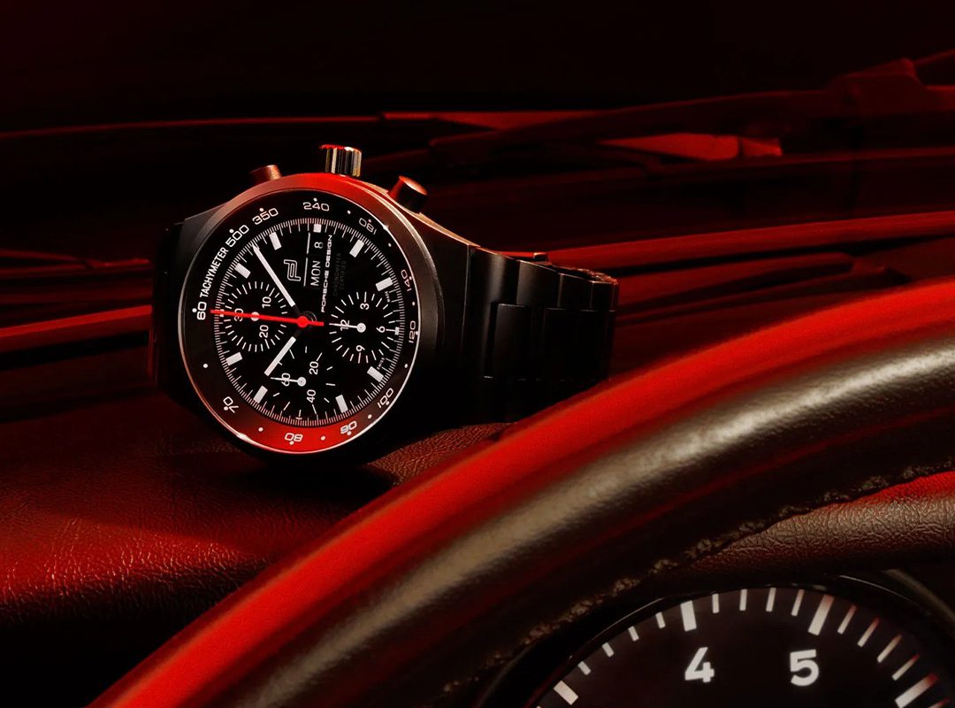 A Porsche Design chronograph wristwatch with a red-accented bezel rests on a car dashboard, with the steering wheel and speedometer partially visible in the foreground.