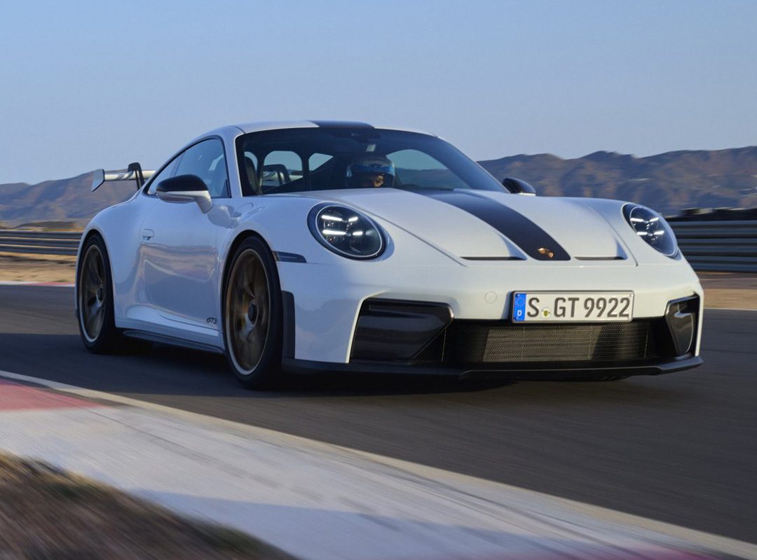 A white Porsche 911 GT3 with a black stripe speeds down the racetrack, mountains towering majestically in the background.