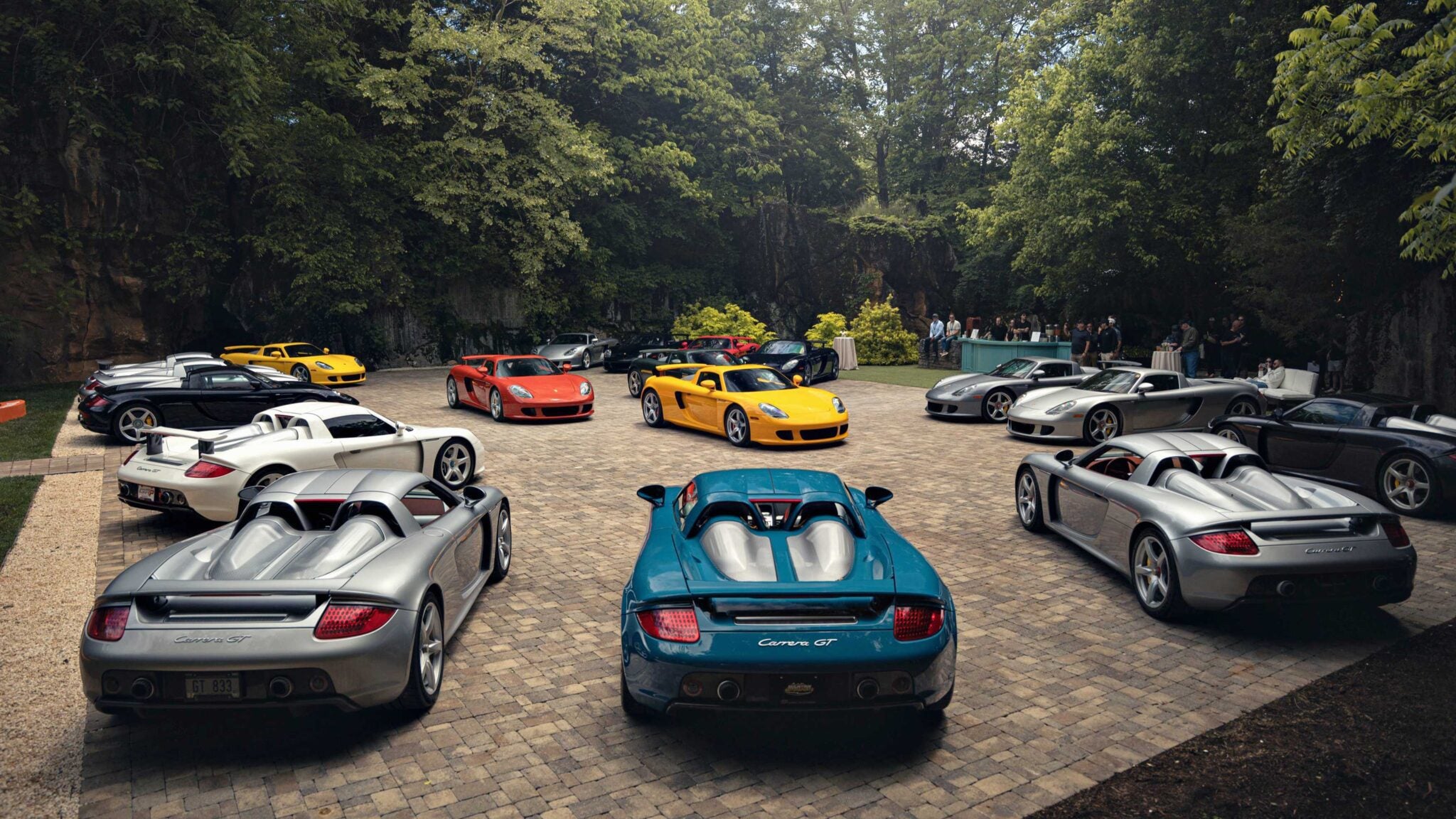 Several Porsche Carrera GT sports cars, part of a 2026 Porsche Plática event, are parked in a circular pattern on a stone courtyard, surrounded by trees and people in the background.