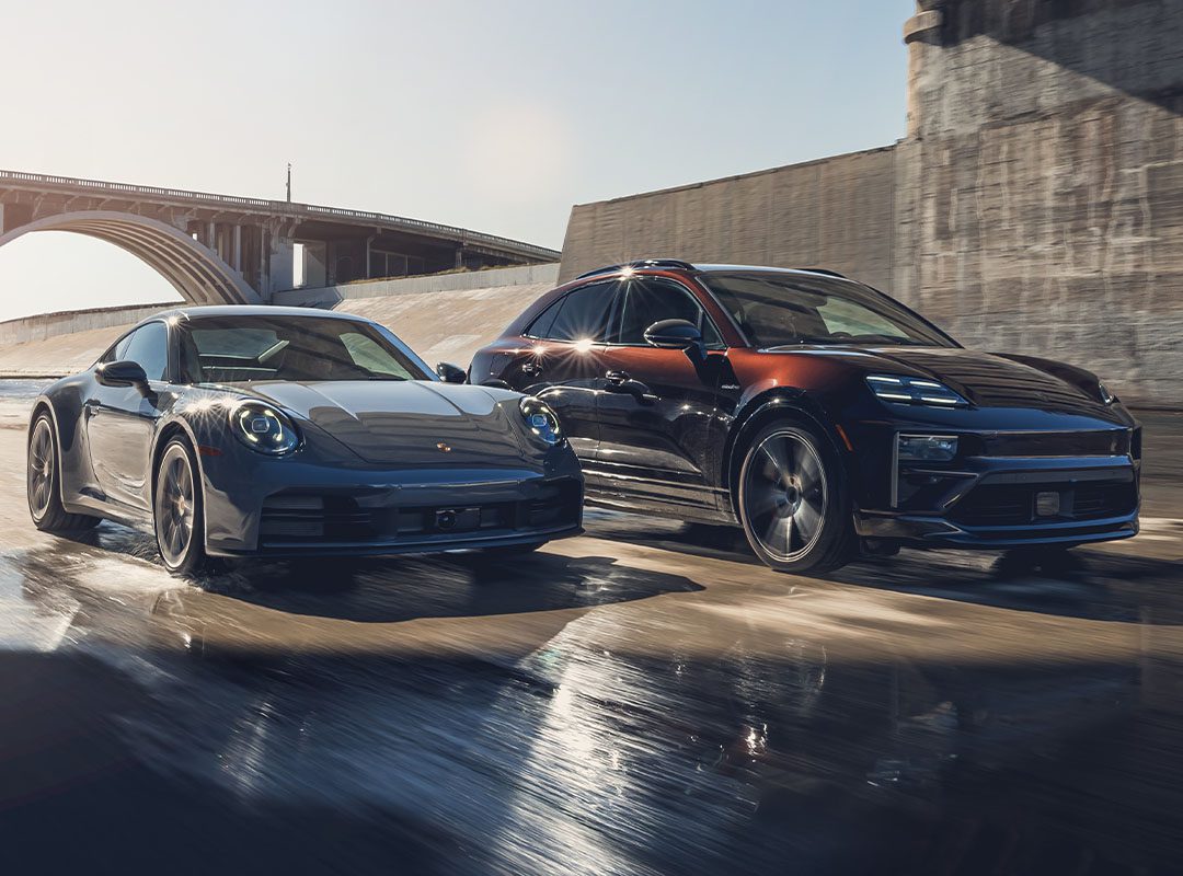 A Porsche sports coupe and an SUV drive side by side on a wet road beside a bridge, reflecting sunlight.