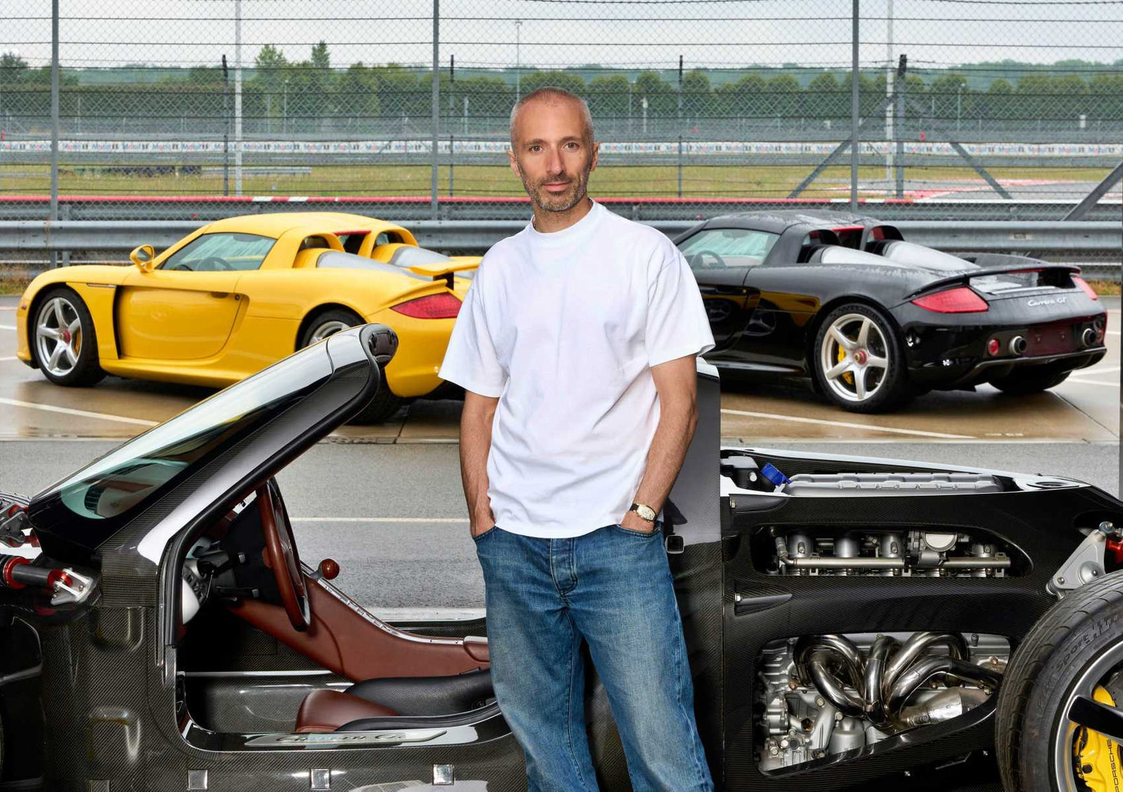 A man in a white t-shirt and jeans stands in front of three high-performance Porsche sports cars, including a Carrera GT, on a racetrack with a chain-link fence—capturing the essence of speed and style like a tribute capsule collection.