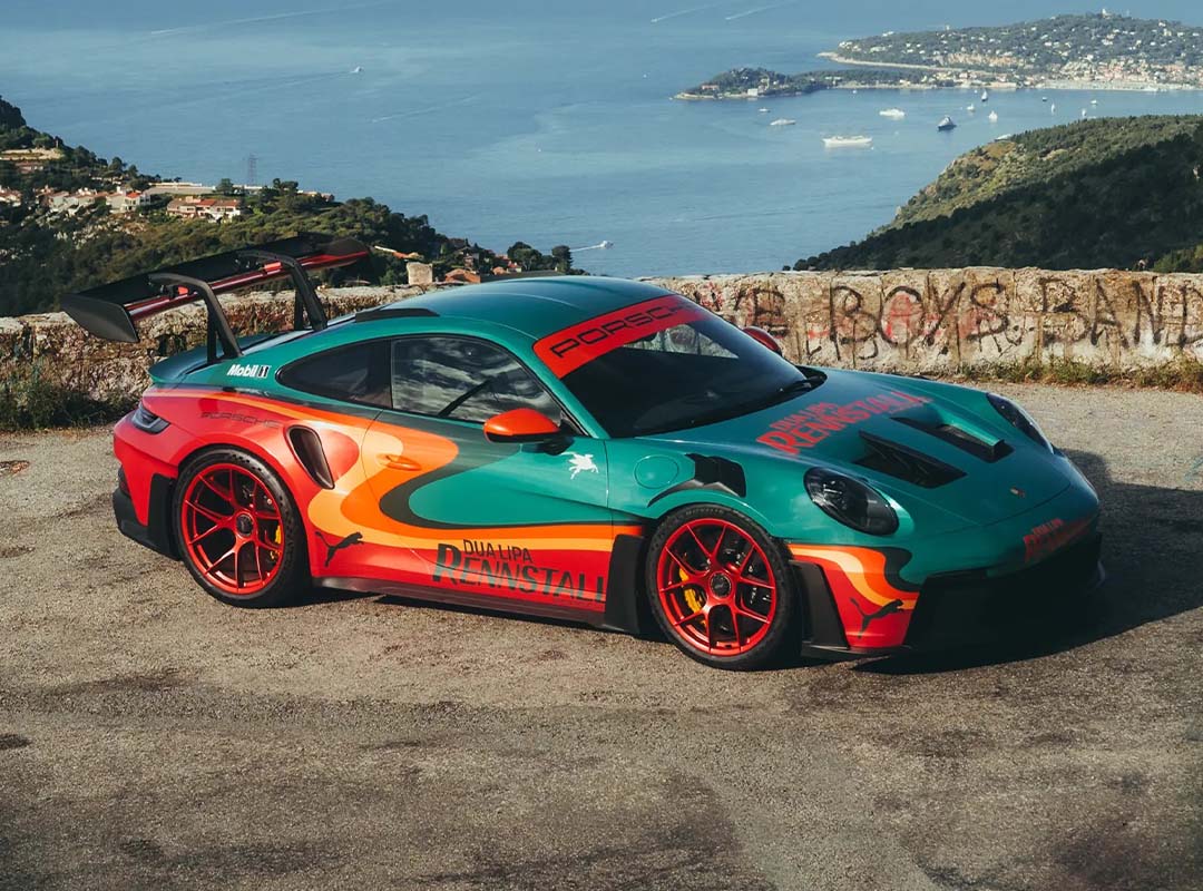 A green and orange Porsche 911 GT3 RS with a rear wing is parked on a scenic overlook, with a coastal town and sea in the background—looking like it could headline an RM Sotheby's Auction or feature in Dua Lipa's next music video.
